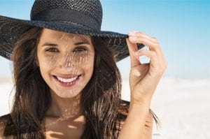 Woman smiling on the beach, wearing a black hat to protect her from the sun.